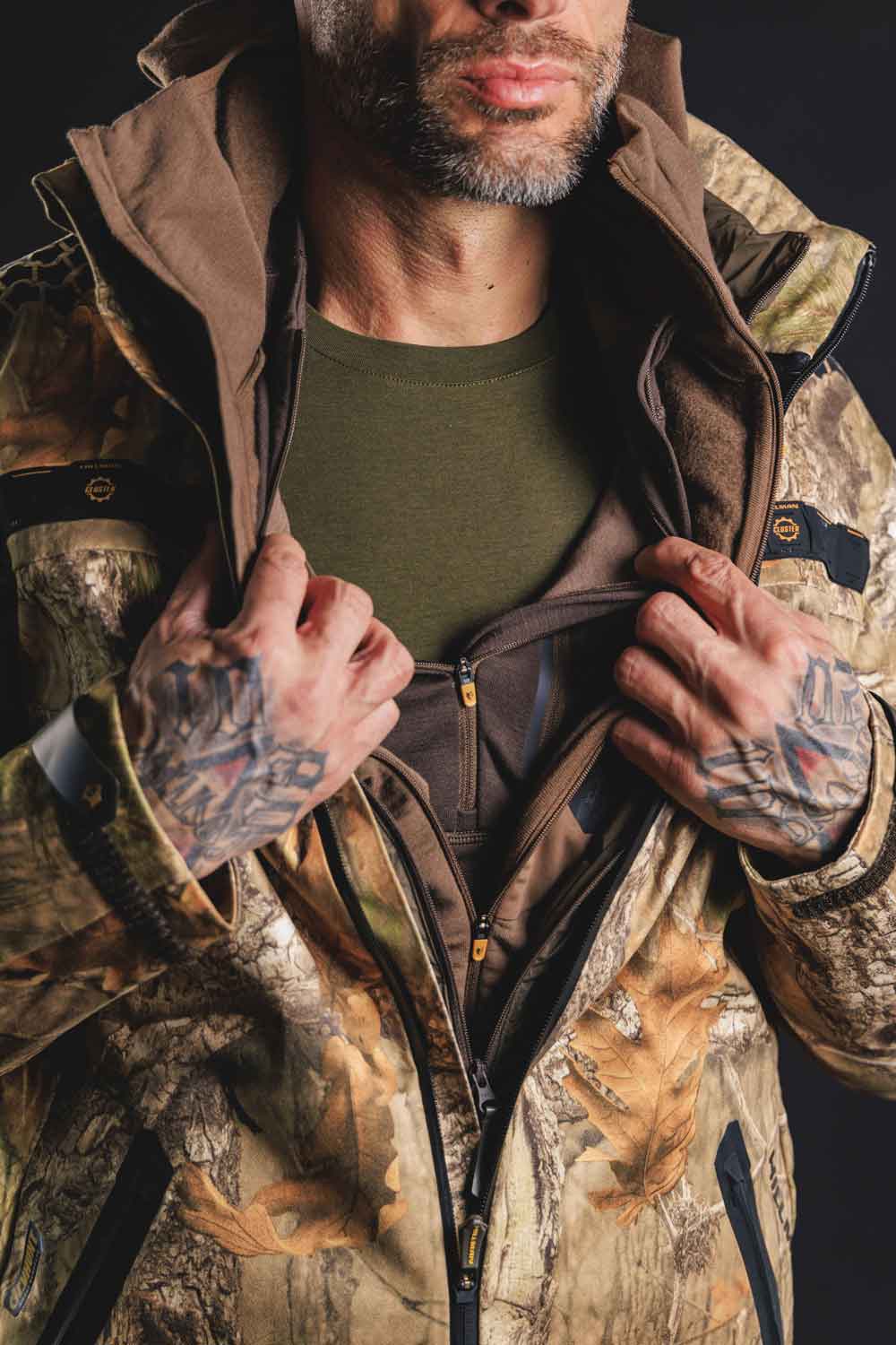 A person wearing a camo-patterned jacket and green shirt is adjusting or zipping up their clothing. They have tattooed hands and a beard. The background is dark, emphasizing the outdoor gear.