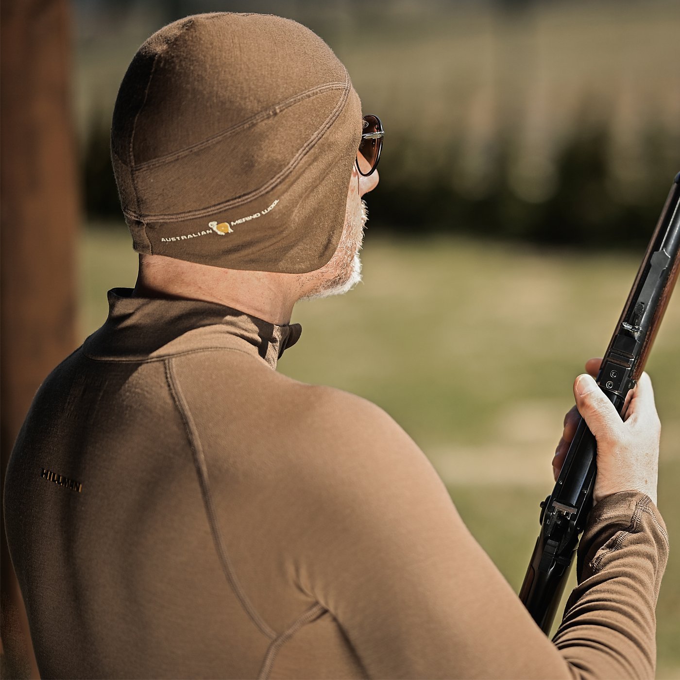 A back-view of a man wearing a brown merino beanie and matching base layer while holding a rifle. This high-quality hunting beanie is crafted from Australian merino wool for superior warmth in the field.