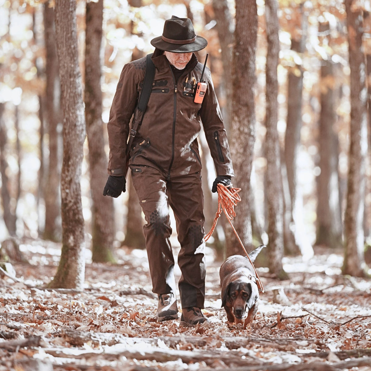 Man in cold weather hunting pants and a wide-brimmed hat leading a tracking dog along a leaf-covered forest path with light frost on the ground