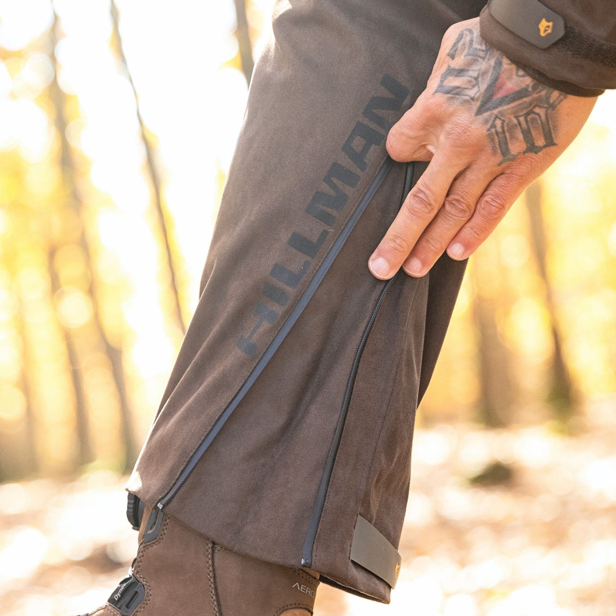 A person with a tattooed hand unzips the side of Hillman's Insulated Hunting Pants for winter while standing in a sunlit forest, wearing brown boots.