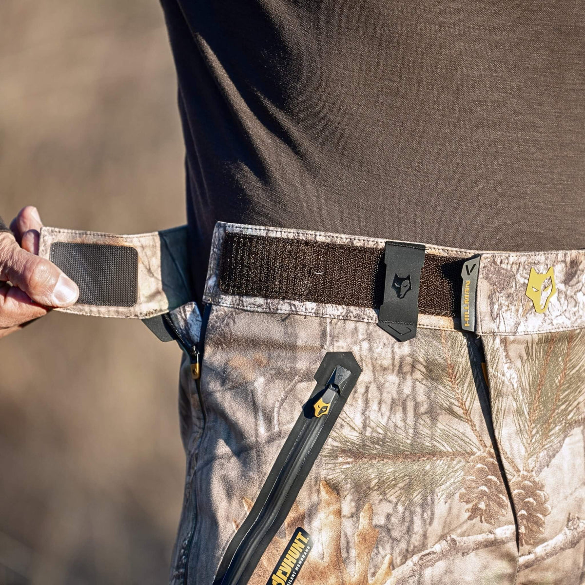 Close-up of Hillman camo hunting pants showing the velcro waistband being adjusted, with branded clips and zip pocket visible. Waterproof camo hunting pants with a silent, adjustable fit system.