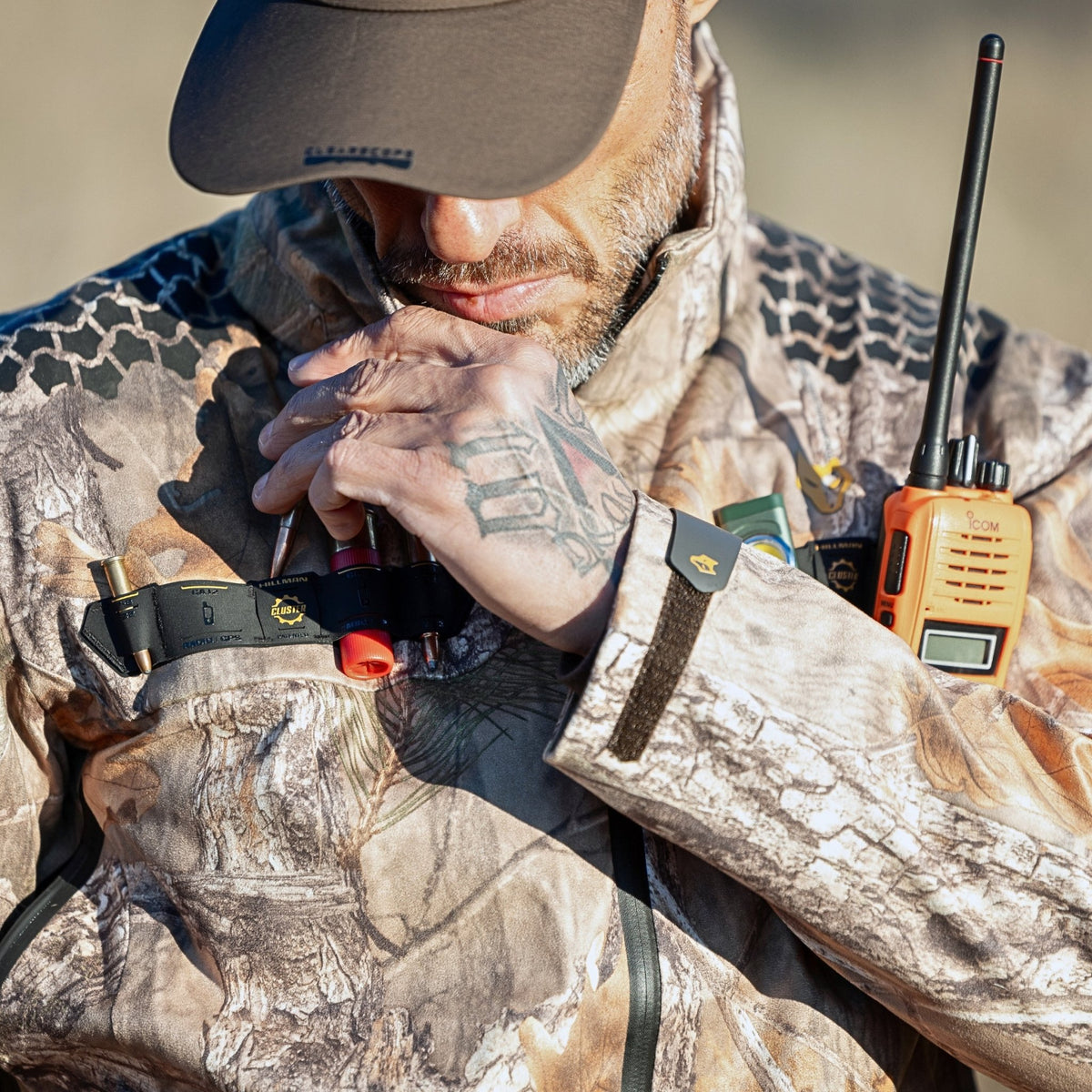 Detail shot of a hunter in a Hillman camo jacket with a fully loaded chest cluster, radio and cartridges to hand. A camo rain jacket built around the idea that every second of access counts in the field.
