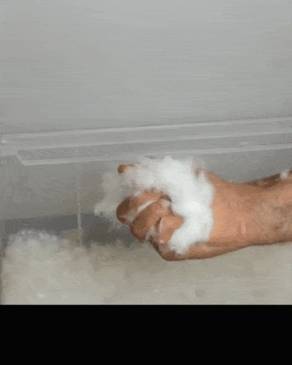 A hand squeezes a clump of wet cotton-like material, releasing droplets of water, with more of the same material spread out below.