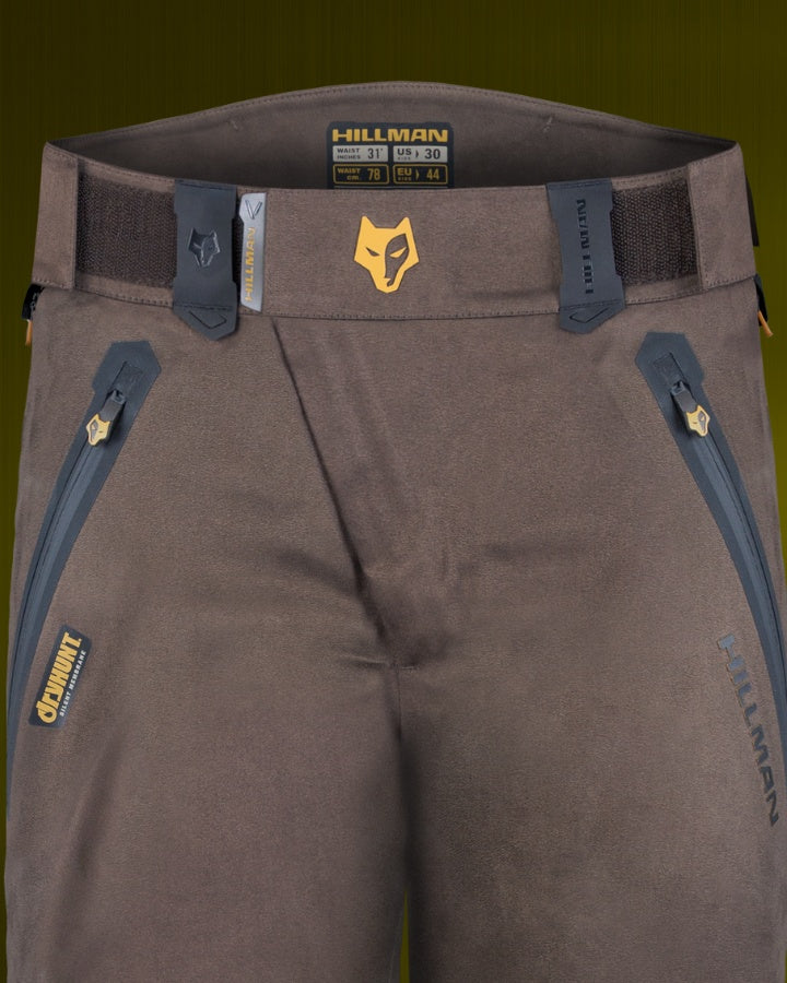 Close-up of brown pants with zipper pockets and a belt. The waistband features a logo of a fox head and the brand name HILLMAN. The pants have decorative stitching and are positioned against a green background.