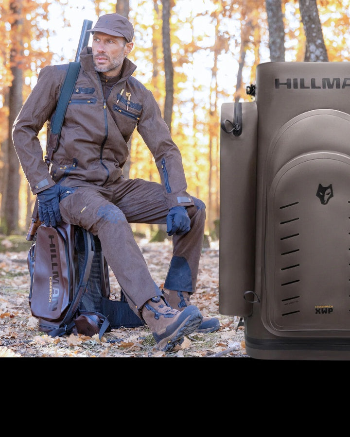 A person wearing brown outdoor hunting gear and a hat is crouched in a forest with fallen leaves. They have a backpack and a shotgun slung over their shoulder. A Hillman-branded case is in the foreground.