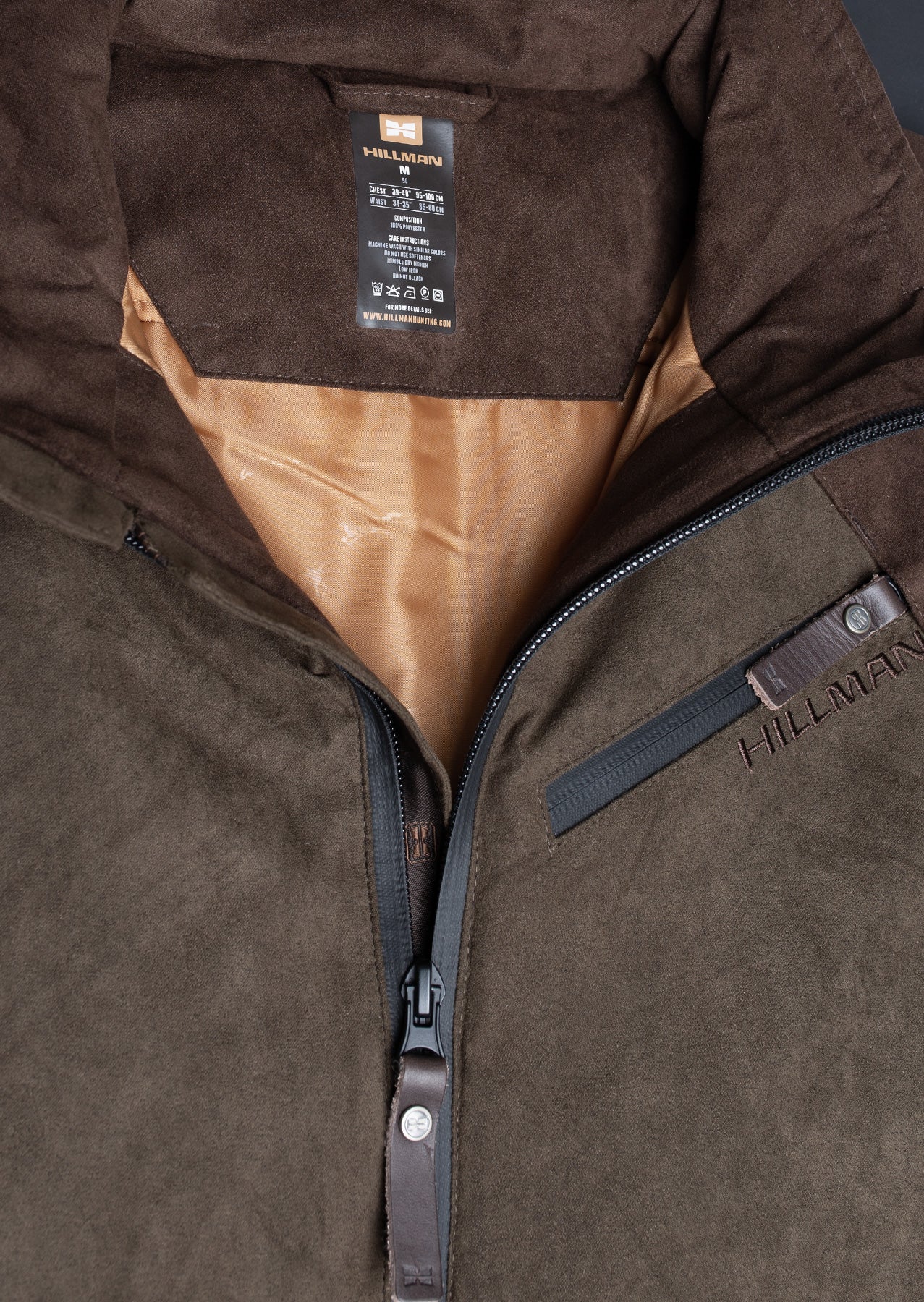 Close-up of the Fusion Hunting Jacket in brown suede, featuring a lightweight design, partially open zipper showing a tan lining, HILLMAN brand label, and an embroidered logo on the chest pocket flap.