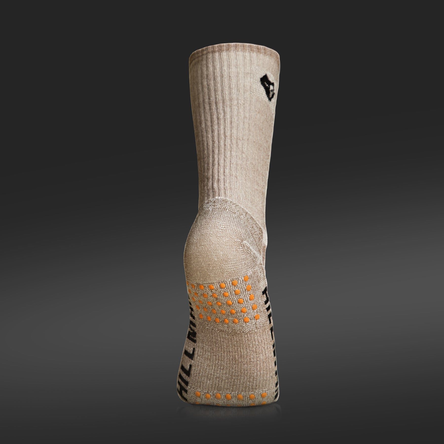 A close-up, side-angle view of a beige merino hunting sock standing upright against a dark grey background. The sock features a ribbed knit texture, a reinforced heel, and bright orange silicone grip dots on the sole for added traction.