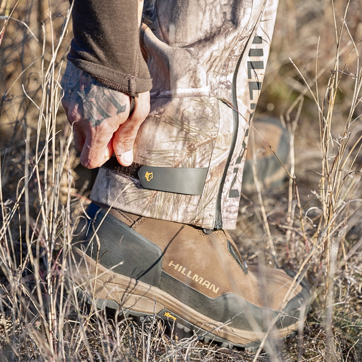 Ankle detail of Hillman camo hunting pants being secured over a boot in dry grassland. The close-fitting zip and hook system reflects the lightweight waterproof build throughout these hunting pants.