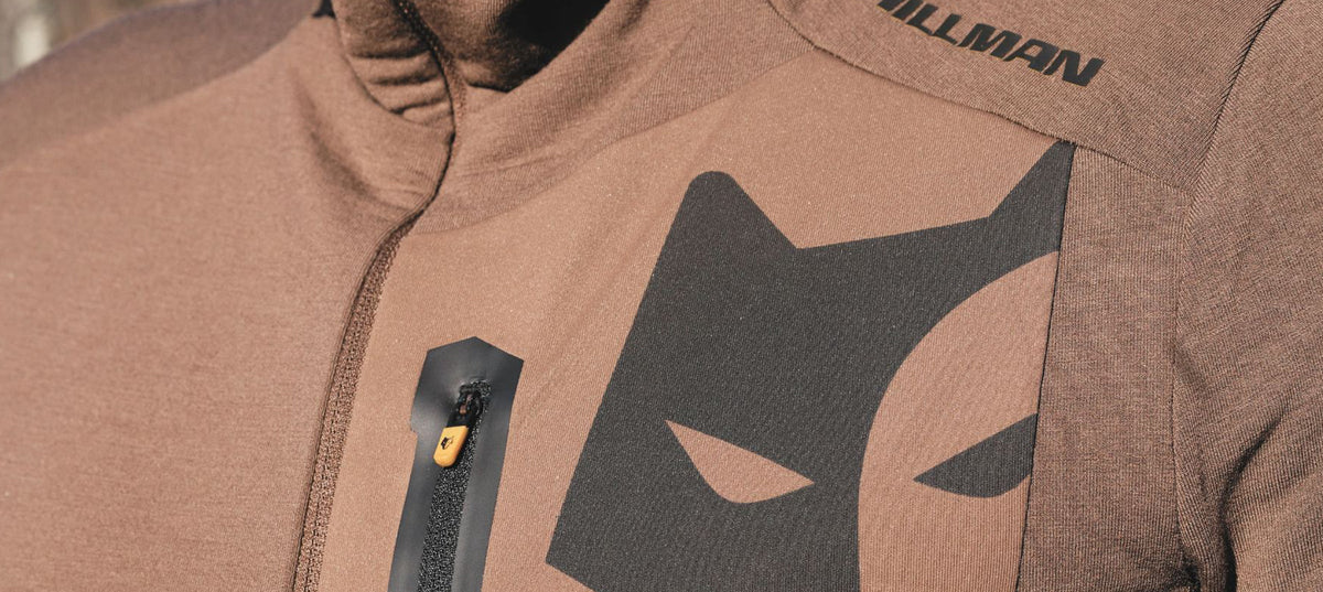 Close-up of a brown jacket featuring a black stylized animal face logo and a small zippered chest pocket with a yellow zipper pull. The word HILLMAN is printed near the shoulder.