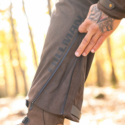 A person with a tattooed hand unzips the side of Hillman Cold Weather Camo Pants - Winter Hunting AlphaShell | 6OL while standing in a sunlit forest, wearing brown boots.