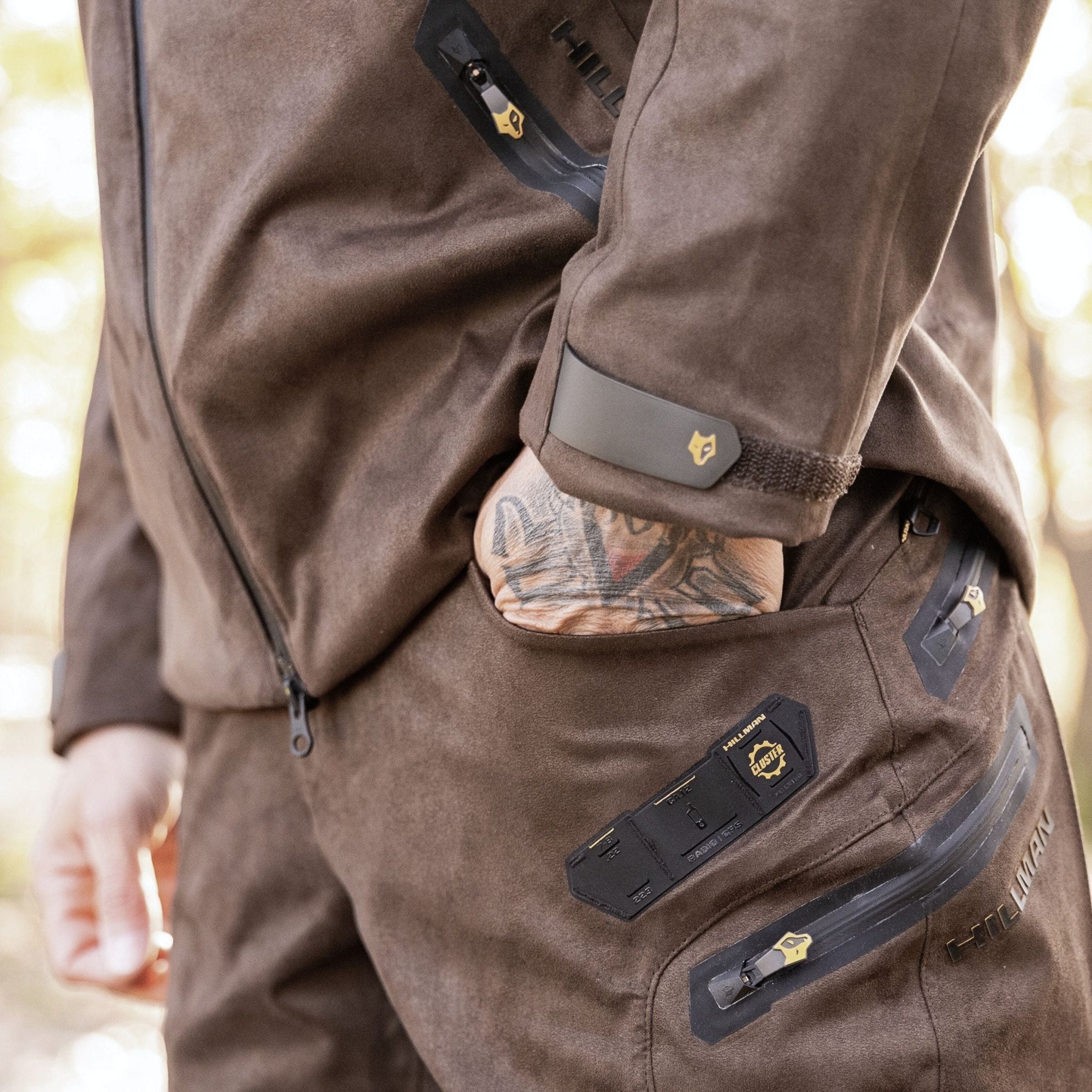 A person wearing Hillman Cold Weather Camo Pants – Winter Hunting AlphaShell | 6OL stands outdoors. Their tattooed hand rests in a pocket. The pants and a brown jacket have multiple zippers, small yellow logos, and black accents.