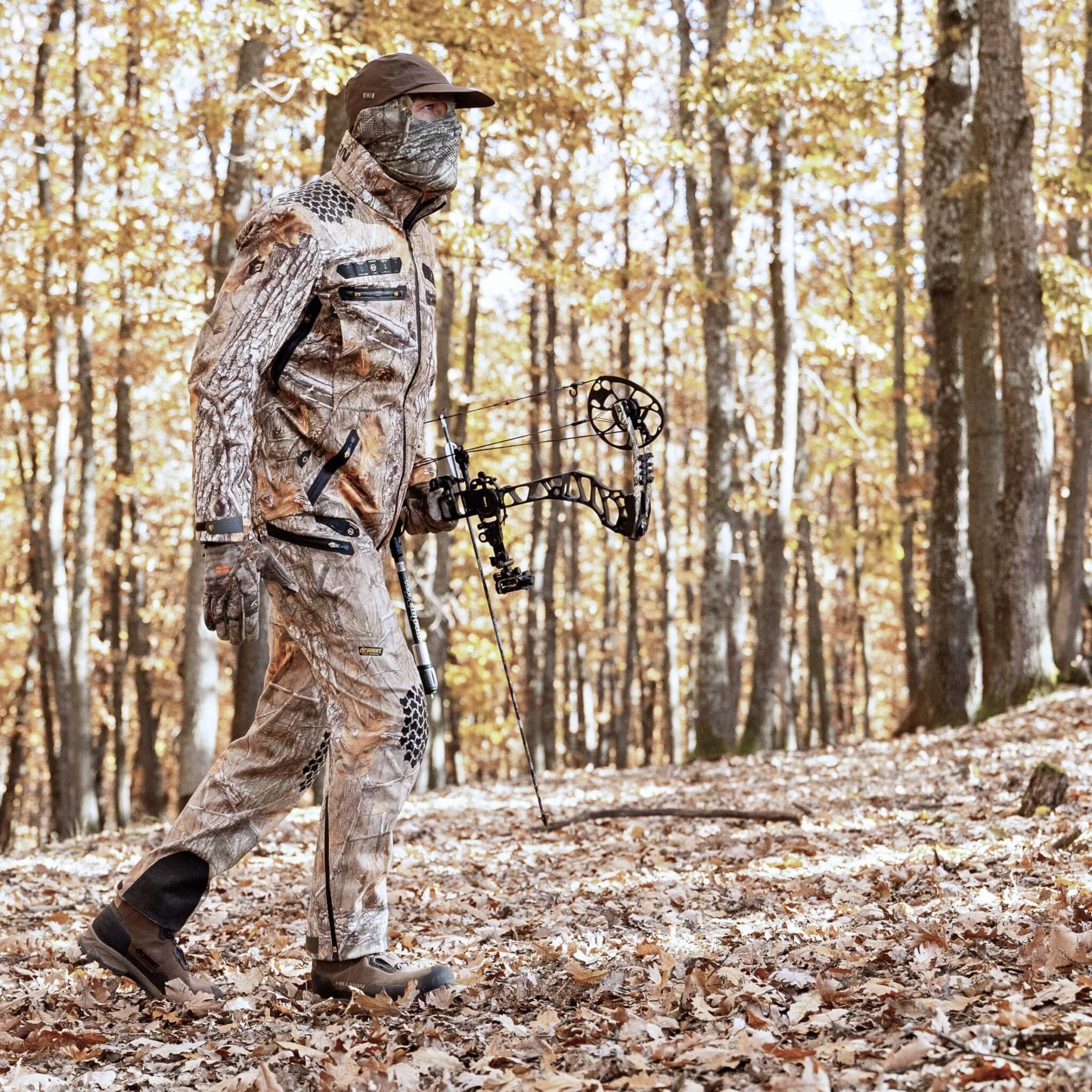 Wearing Hillman’s Cold Weather Camo Pants - Winter Hunting AlphaShell | 6OL, a hunter walks through a sunlit autumn forest with a compound bow, surrounded by fallen leaves and trees.
