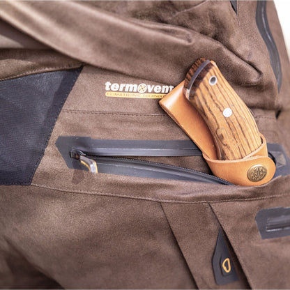 A close-up shows the Hillman Cold Weather Camo Pants - Winter Hunting AlphaShell | 6OL, featuring a brown zippered pocket with a wooden-handled knife in a tan sheath inside, and visible Hillman branding highlights their quality insulation.