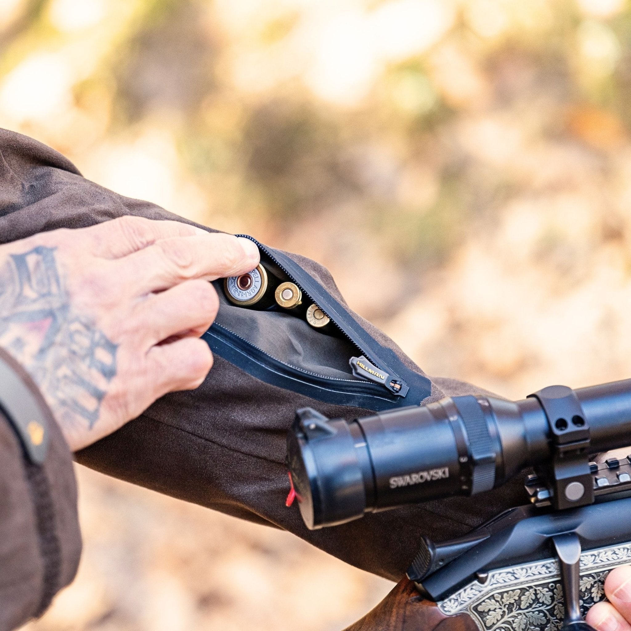 A tattooed hand opens a zippered pocket on the brown sleeve of a Hillman Insulated Hunting Jacket Cold Weather Winter ProShell | 6OL, revealing rifle cartridges inside. A rifle with a Swarovski scope is nearby in the blurred background.