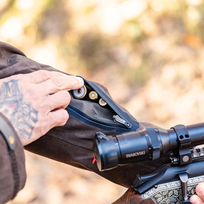 A tattooed hand opens a zippered pocket on the brown sleeve of a Hillman Insulated Hunting Jacket Cold Weather Winter ProShell | 6OL, revealing rifle cartridges inside. A rifle with a Swarovski scope is nearby in the blurred background.