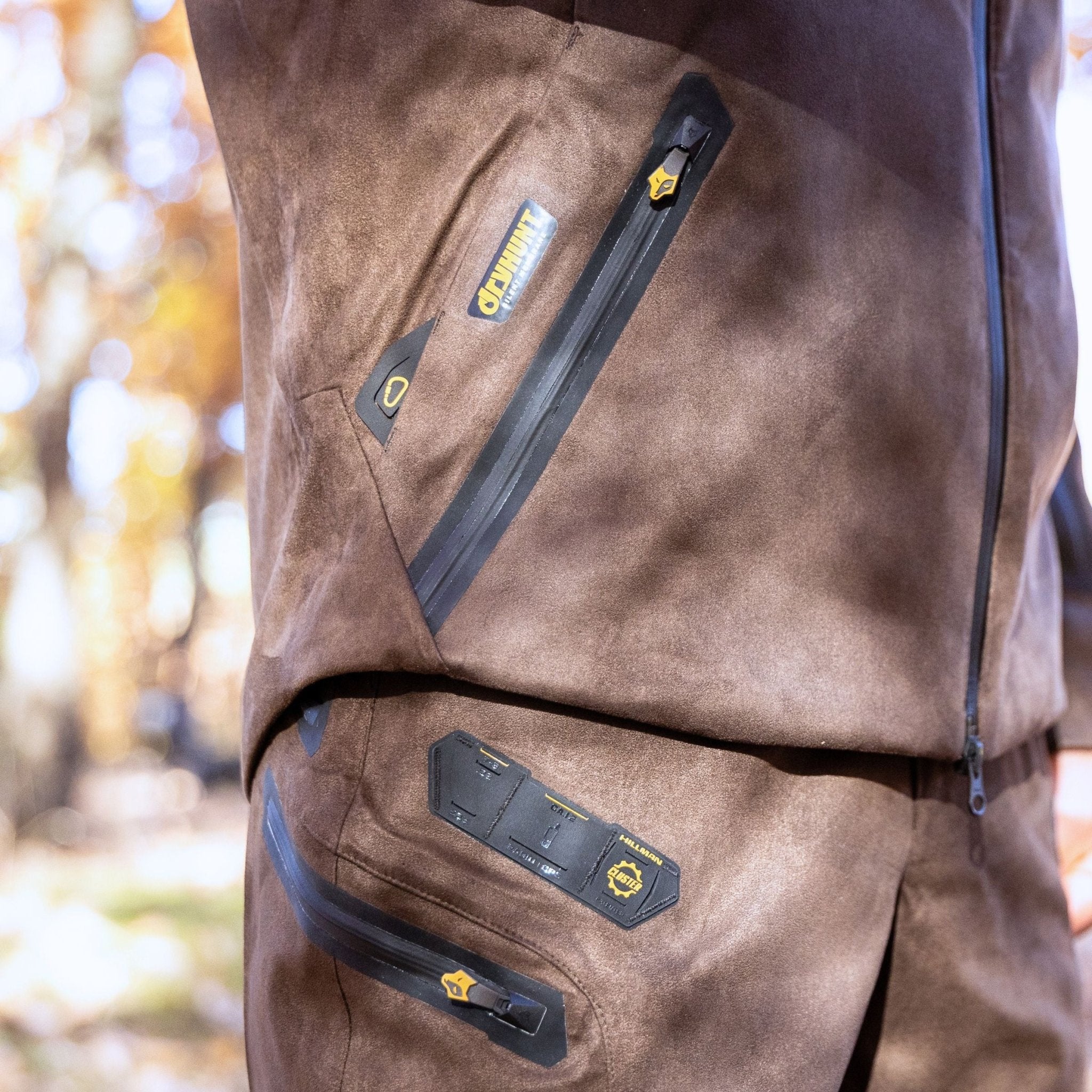 Close-up of a person outdoors in the Hillman 6OL Cold Weather Hunting Jacket, featuring brown fabric, black zippers, and yellow logo details, with blurred trees and sunlight in the background.