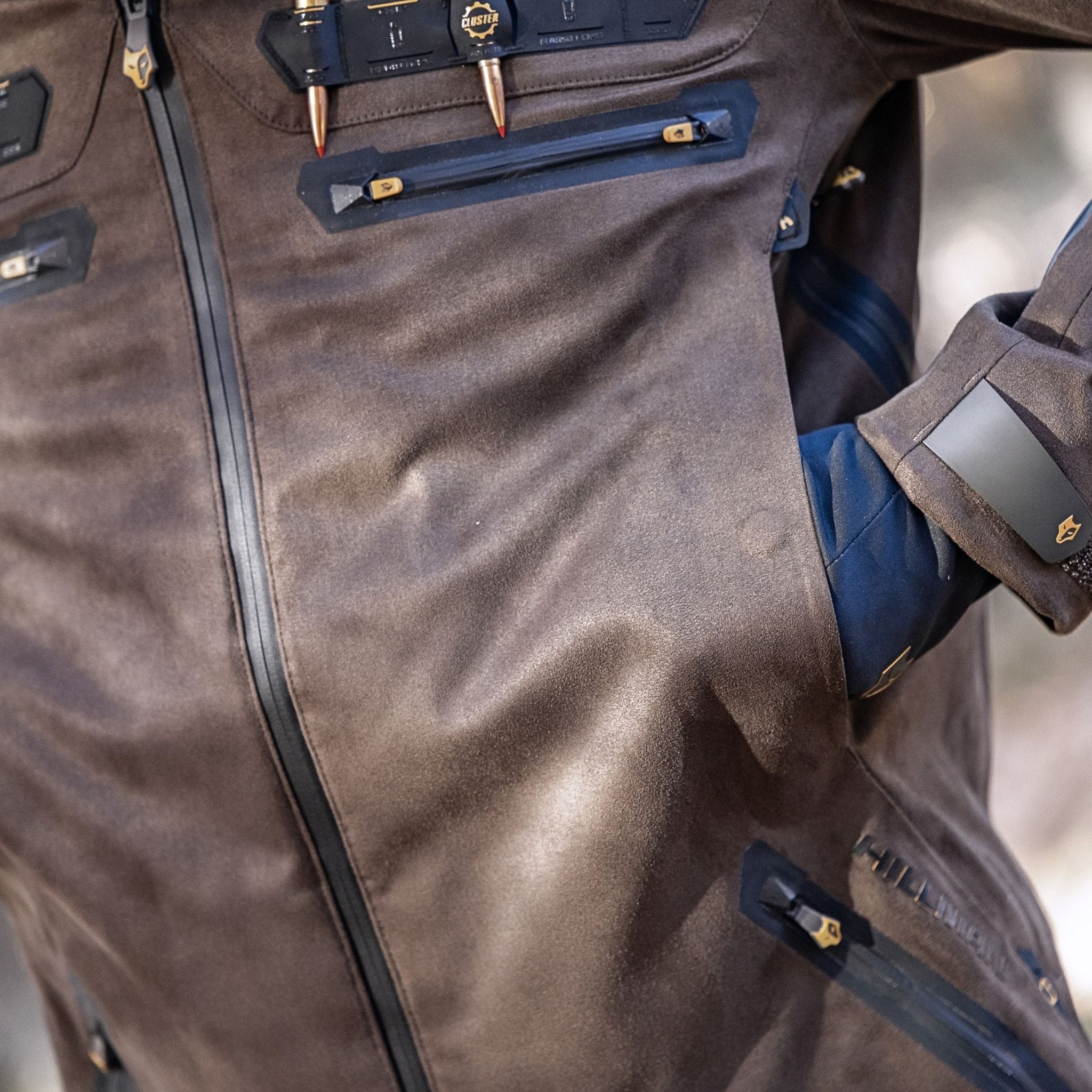A close-up of the Hillman 6OL Cold Weather Hunting Jacket, the warmest insulated coat, shows brown fabric with black zippers and gold accents, partially unzipped to reveal a gloved hand in the pocket and unique fasteners.