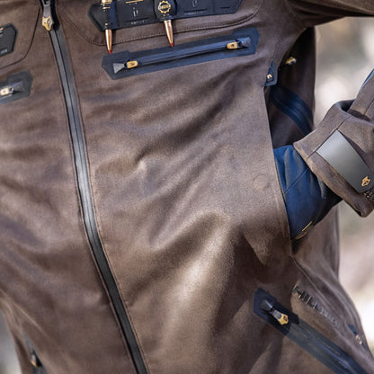 A close-up of the Hillman 6OL Cold Weather Hunting Jacket, the warmest insulated coat, shows brown fabric with black zippers and gold accents, partially unzipped to reveal a gloved hand in the pocket and unique fasteners.
