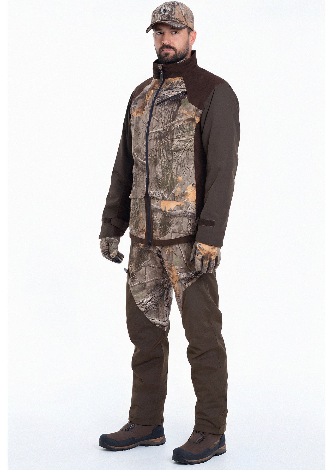 A man with a neutral expression stands facing forward, wearing a Hillman Fusion Hunting Jacket 3DX, camouflage pants, a baseball cap, and brown boots. The outfit suits outdoor or hunting activities. The background is plain white.