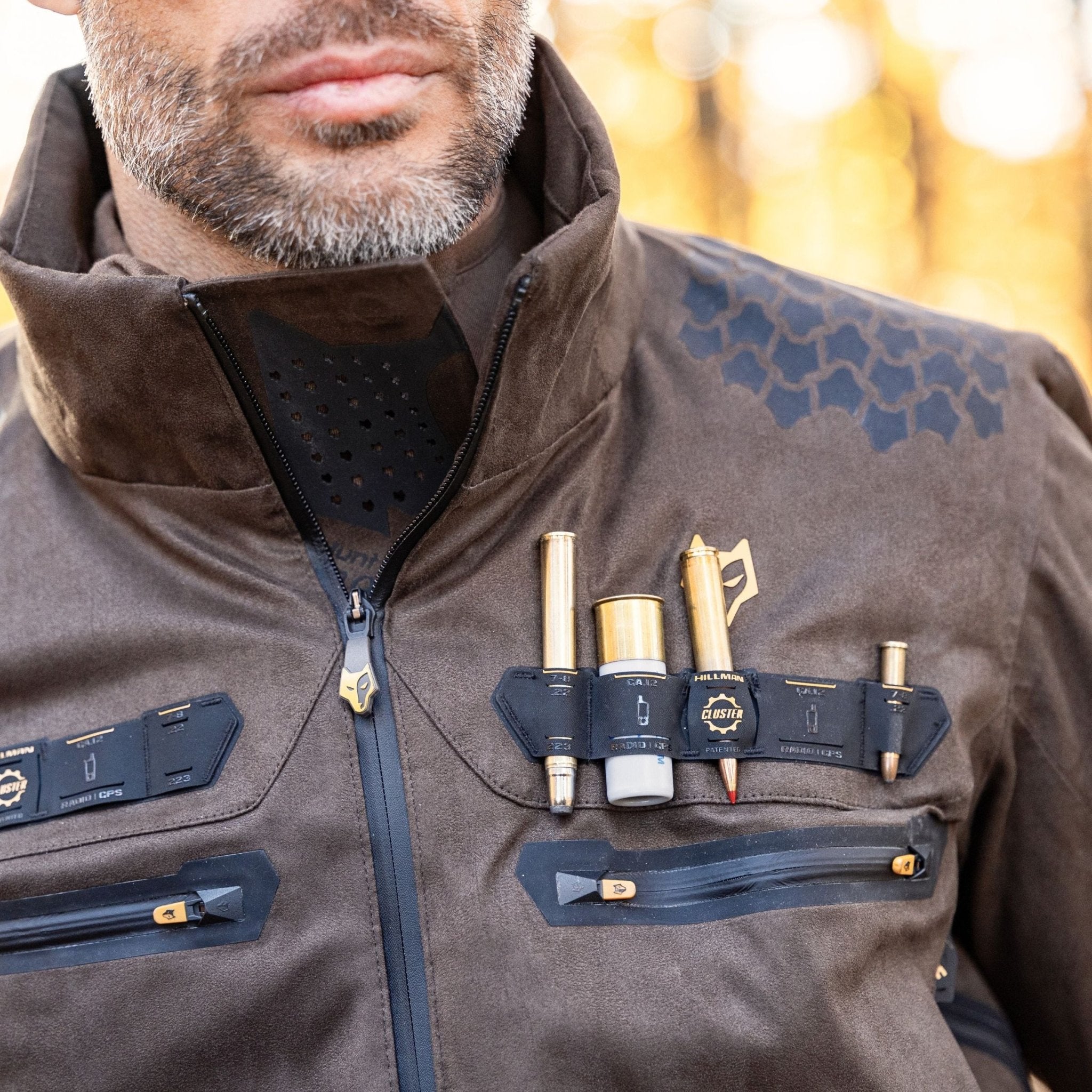 A man wears the Hillman 6OL Cold Weather Hunting Jacket, the warmest insulated coat with black hexagon shoulder patterns. Pens and tools are organized in chest holders; his trimmed beard is partly visible—perfect outdoor gear.