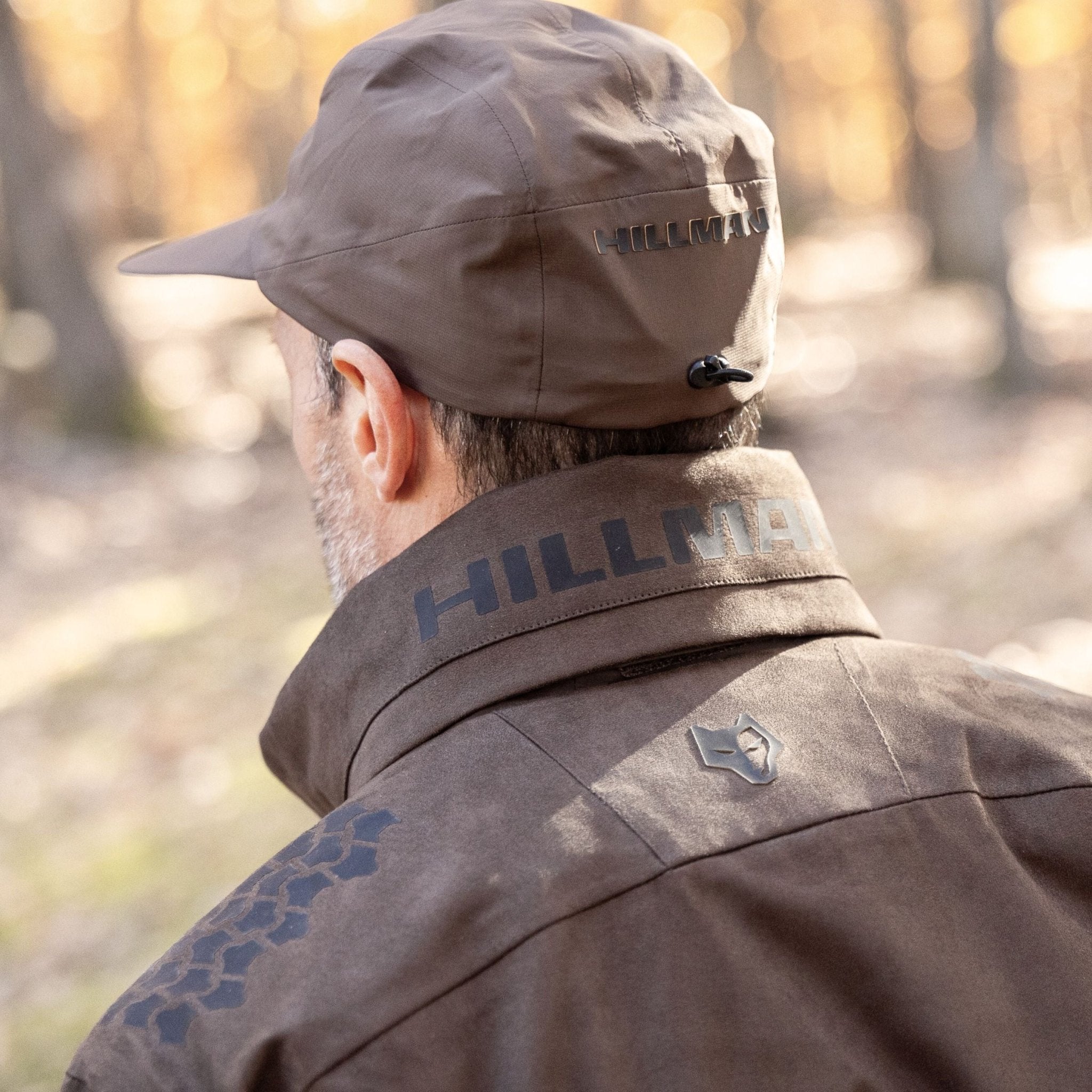 A person in a brown Hillman 6OL Cold Weather Hunting Jacket, the warmest insulated coat, stands outdoors facing trees and sunlight. The photo, taken from behind, showcases this essential gear for nature lovers.