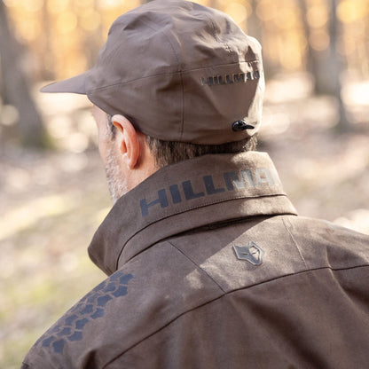 A person in a brown Hillman 6OL Cold Weather Hunting Jacket, the warmest insulated coat, stands outdoors facing trees and sunlight. The photo, taken from behind, showcases this essential gear for nature lovers.